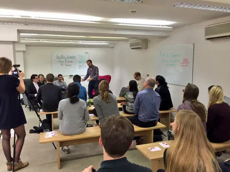 Classroom photo of an audience seated at desks watching a presenter by a whiteboard.