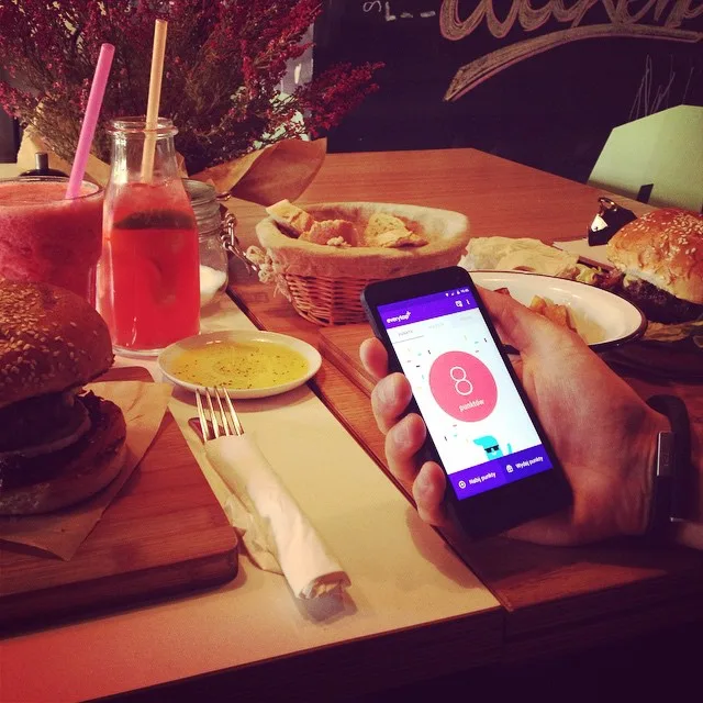 Hand holding a phone showing a pink app screen on a bar table with drinks.