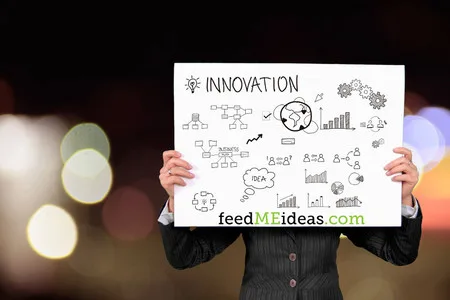 Person in a suit holding a sign with 'INNOVATION' doodles and 'feedMEideas.com' against bokeh lights.