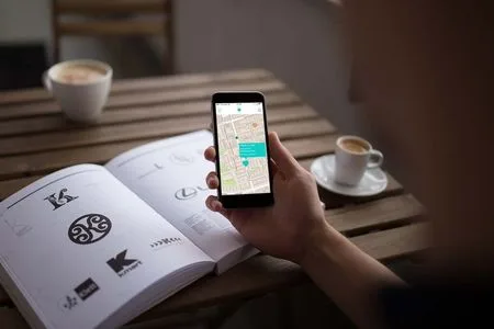 A hand holding a phone showing a map app, on a cafe table with coffee cups and an open book.